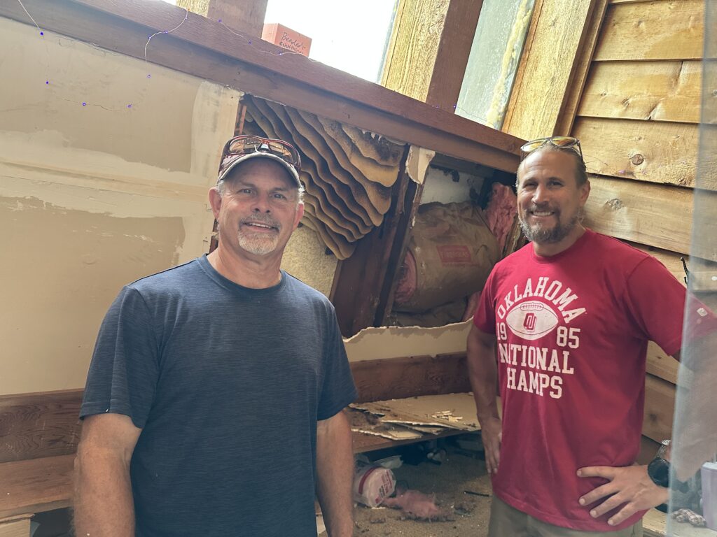 Two men smiling indoors near a wooden display and window.
