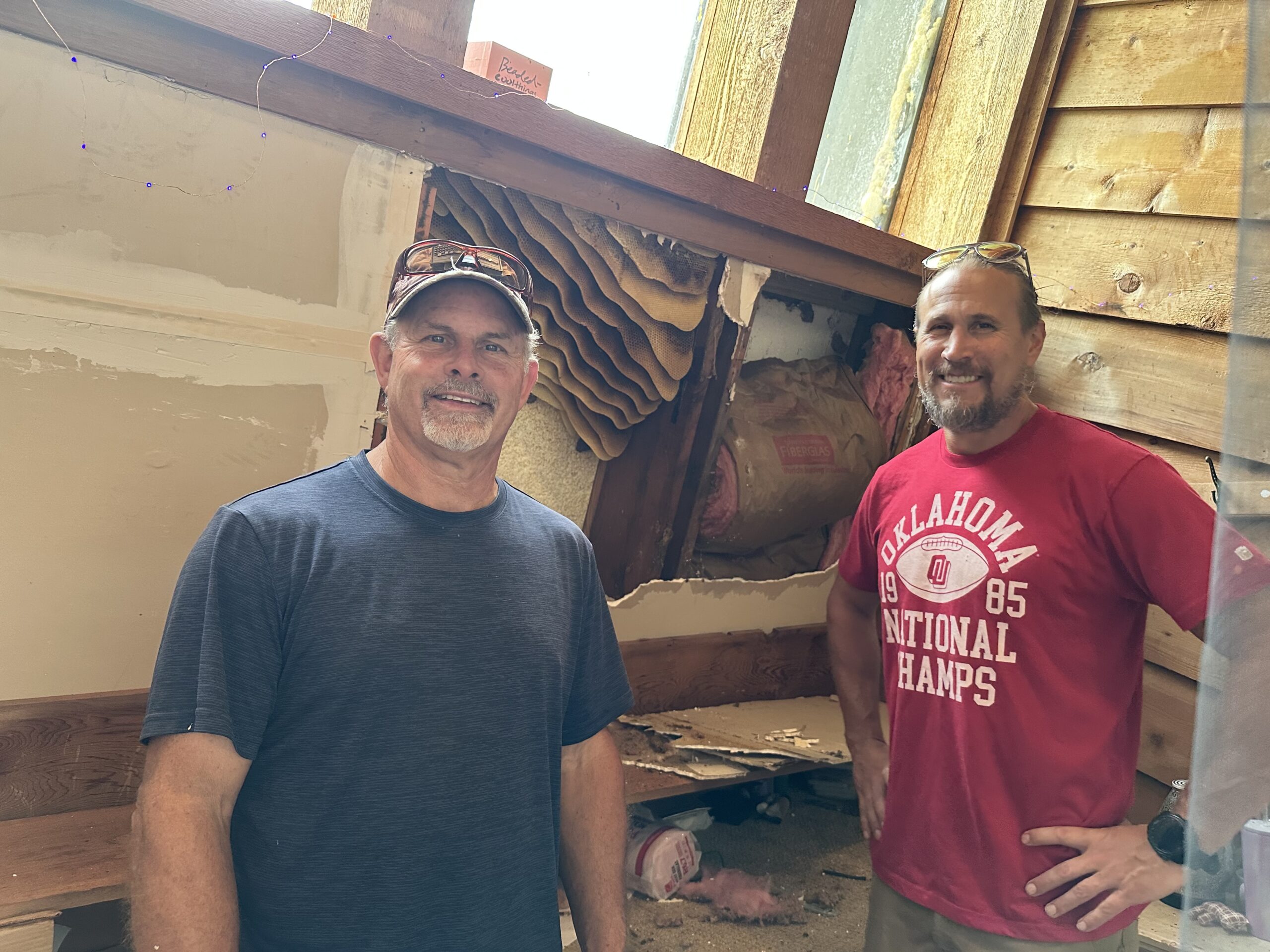 Two men smiling indoors near a wooden display and window.