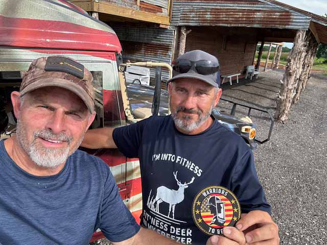 Two men smiling outdoors, one holding a patch with a deer design.